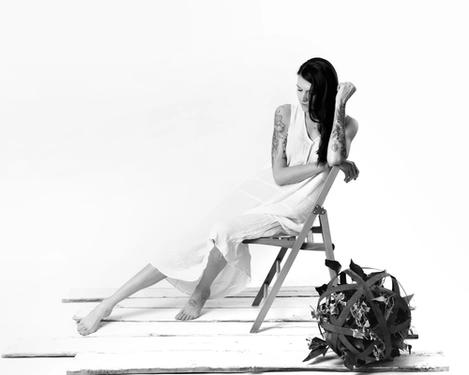 Dark haired woman dressed in white sits on a folding chair looking at the floor