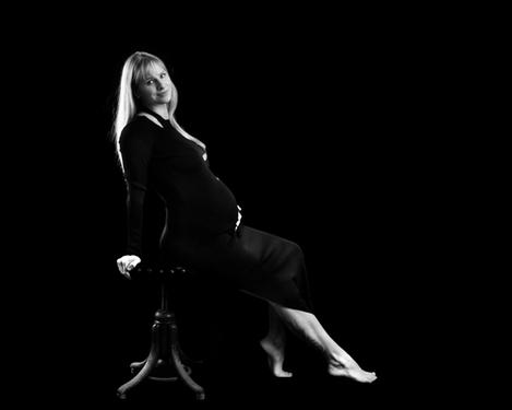 Blonde schwangere Frau in elegantem schwarzen Kleid sitzt seitlich auf einem Hocker und blickt in die Kamera; Schwarzer Hintergrund, sw-Bild