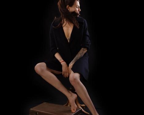 Dark-haired woman dressed in black sits on a stool looking to the side, black background