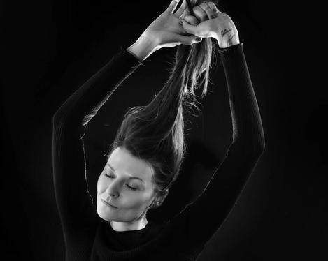 Dark-haired woman in black bodysuit sits on a chair with closed eyes and pulls up her hair, black background