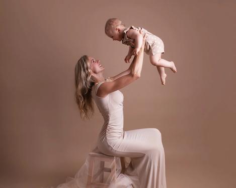 Mutter in bodenlangem Kleid sitzt auf einem Hocker und hält ihr einjähriges Baby in die Höhe.  Beide sind beige gekleidet bei gleichfarbigem Studioset.