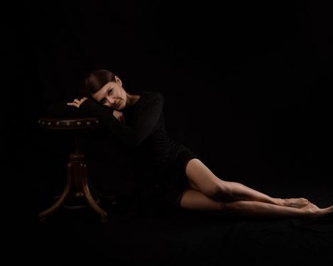 Woman in black dress sits on the floor with her legs stretched out, leaning on a stool. Her gaze is directed towards the camera, black background.
