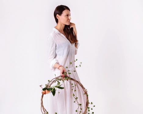 Frau in weißem Kleid hält einen Blumenkranz in der Hand, sie blickt geradeaus