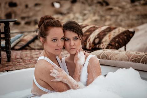 A same-sex female couple sits in an outdoor bathtub with their heads together.