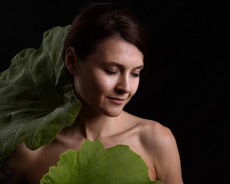 Portait einer dunkelhaarigen Frau, umrahmt von zwei großen grünen Blättern. Ihre Augen sind geschlossen und Kopf ist zum Licht geneigt.