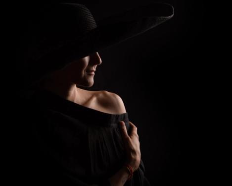 Side view of a woman in a black hat wearing a black dress in pincer light. One shoulder is visible, black background.