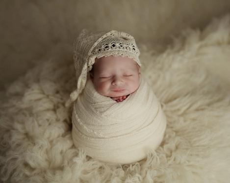 Baby mit Zipfelmütze eingewickelt in creme farbigen Tuch auf fluffigem Schaffell