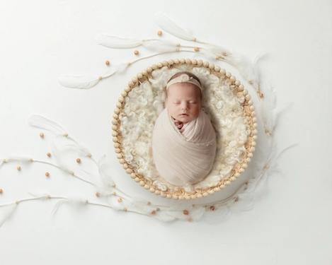 Eingewickeltes Neugeborenes in rundem, mit Fleece ausgepolstertes Nesterl auf weißem Hintergrund. Dekoriert mit weißen Federn