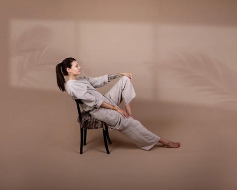 Dark-haired woman sits on an armchair in beige trousers in a studio set of the same colour. One leg is bent, the other stretched out. Shadows of palm trees can be seen in the background.