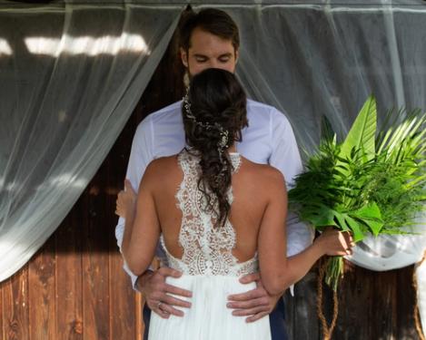 Bride and groom face each other in front of a rustic wooden wall and white curtain. Bride stands with her back to the camera, holding a green bridal bouquet.