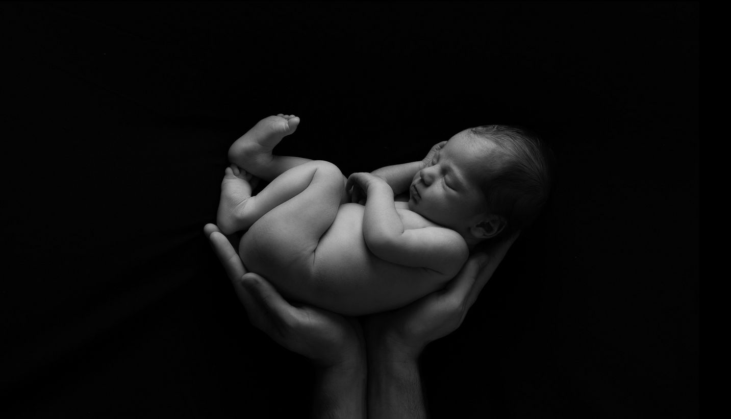 Schlafendes, nacktes Baby in Papas Händen vor schwarzem Hintergrund