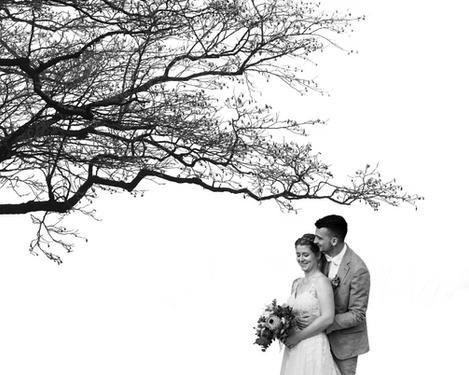 Groom is standing behind the bride, both are smiling and introverted.  A thick branch sticks out into the picture - bw image