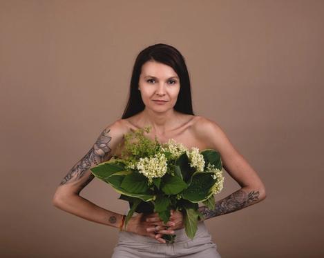 Dark-haired woman holds bouquet of flowers in front of her upper body, her arms are tattooed