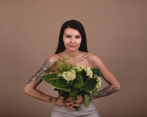 Dunkelhaarige Frau hält Blumenstrauß vor ihrem Oberkörper, ihre Arme sind tätowiert