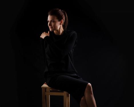 Dark haired woman in black dress sits on a chair facing the side, black background