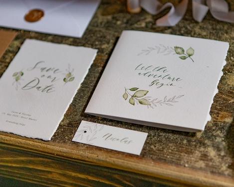 Wedding stationery on a rustic wooden table. Torn handmade paper with olive green leaf decor and script font