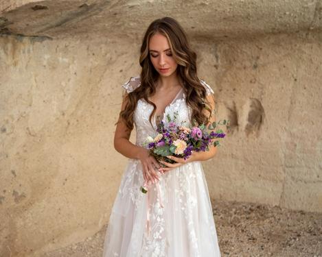Braut steht in einer Sandsteinhöhle und betrachtet ihren rosa/lila Brautstrauß