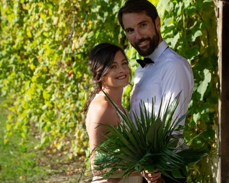 Bride and groom stand together in front of a green leafy background and smile at the camera