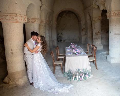 Braut und Bräutigam lehnen an einer Säule in einem sandsteinfarbenen Raum wo auch die Hochzeitstafel steht.