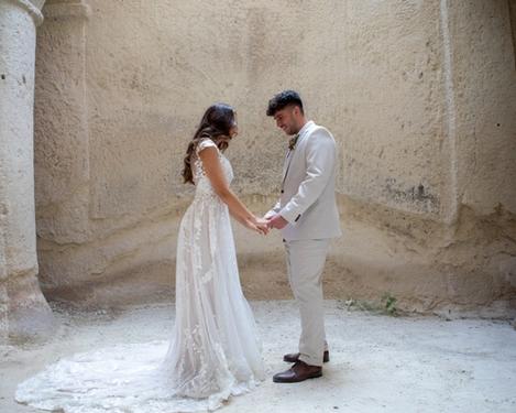 Brautpaar steht einander gegenüber in einem sandsteinfarbenen Raum mit Säulen. Sie halten sich an den Händen und betrachten einander.