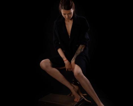 Dark haired woman dressed in black sitting on a stool looking down, black background
