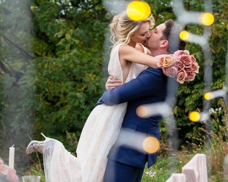 Bräutigam hebt Braut hoch, dabei küssen sie sich. Im Vordergrund der gedeckte Tisch mit Lichtpunkten   