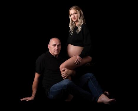 Blonde schwangere Frau mit ihrem Partner, beide in schwarzem Oberteil blicken entspannt zur Camera
