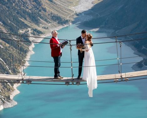 Groom and groom stand on a suspension bridge and kiss, a wedding speaker stands opposite, behind and below them a turquoise lake framed by mountains