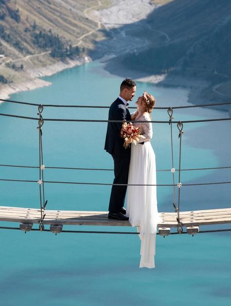 Wedding couple in classic dress facing each other on a suspension bridge in the mountains. In the background are mountains and a turquoise blue reservoir. 