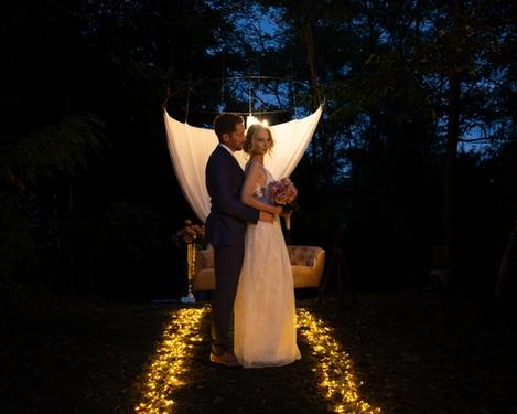 Groom stands behind the bride. She looks at the camera, he at her. Evening atmosphere outdoors with fairy lights on the ground.