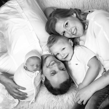 Familie mit Neugeborenem und Kleinkind liegen auf einem weißen Fell und lächeln in die Kamera