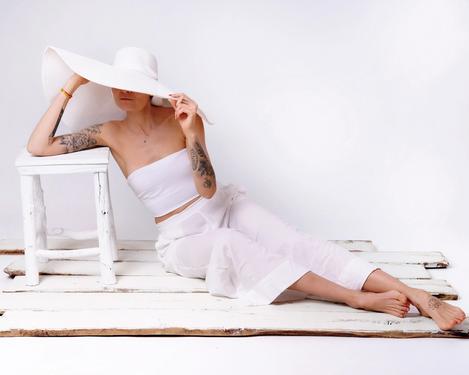 Weiß gekleidete Frau mit weißem Hut sitzt auf Holzbrettern, ihr Gesicht ist von der Hutkrempe verdeckt