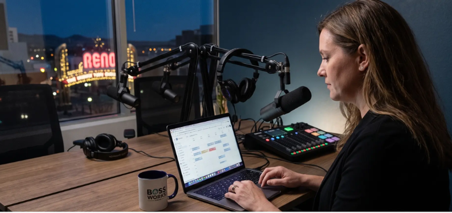 A woman reviews a calendar on her laptop in a podcast studio with the Reno sign visible outside
