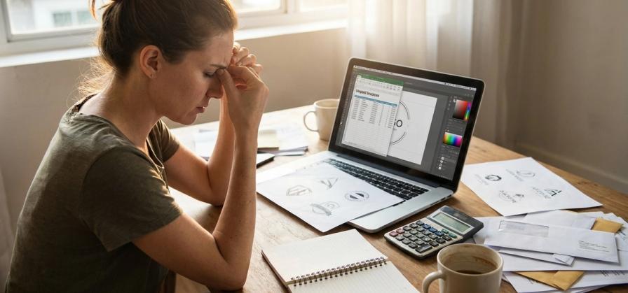 A stressed woman sits at a desk with a laptop, calculator, and scattered papers