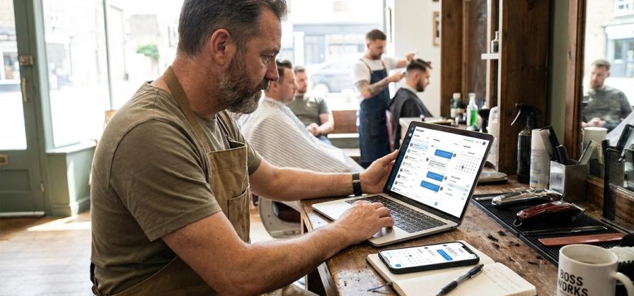 A bearded barber in a brown apron uses a laptop showing a calendar at a barbershop counter