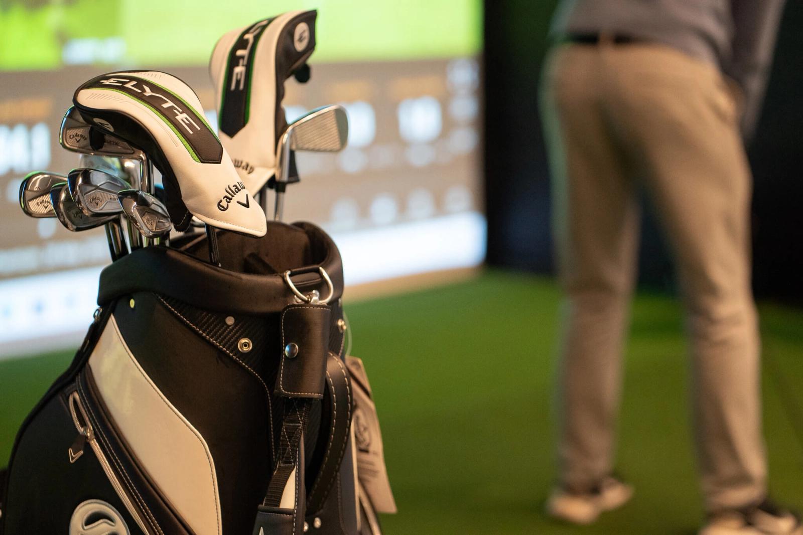 A close-up of a golf bag filled with clubs in the foreground, while a person in gray pants prepares to swing at a golf simulator screen—a perfect setting for golf lessons in London.