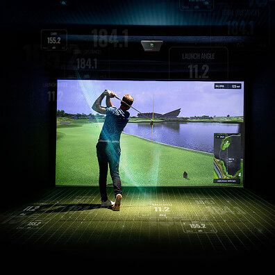 A person swings a golf club in an indoor golf simulator, facing a large screen displaying a realistic golf course scene with stats and metrics.
