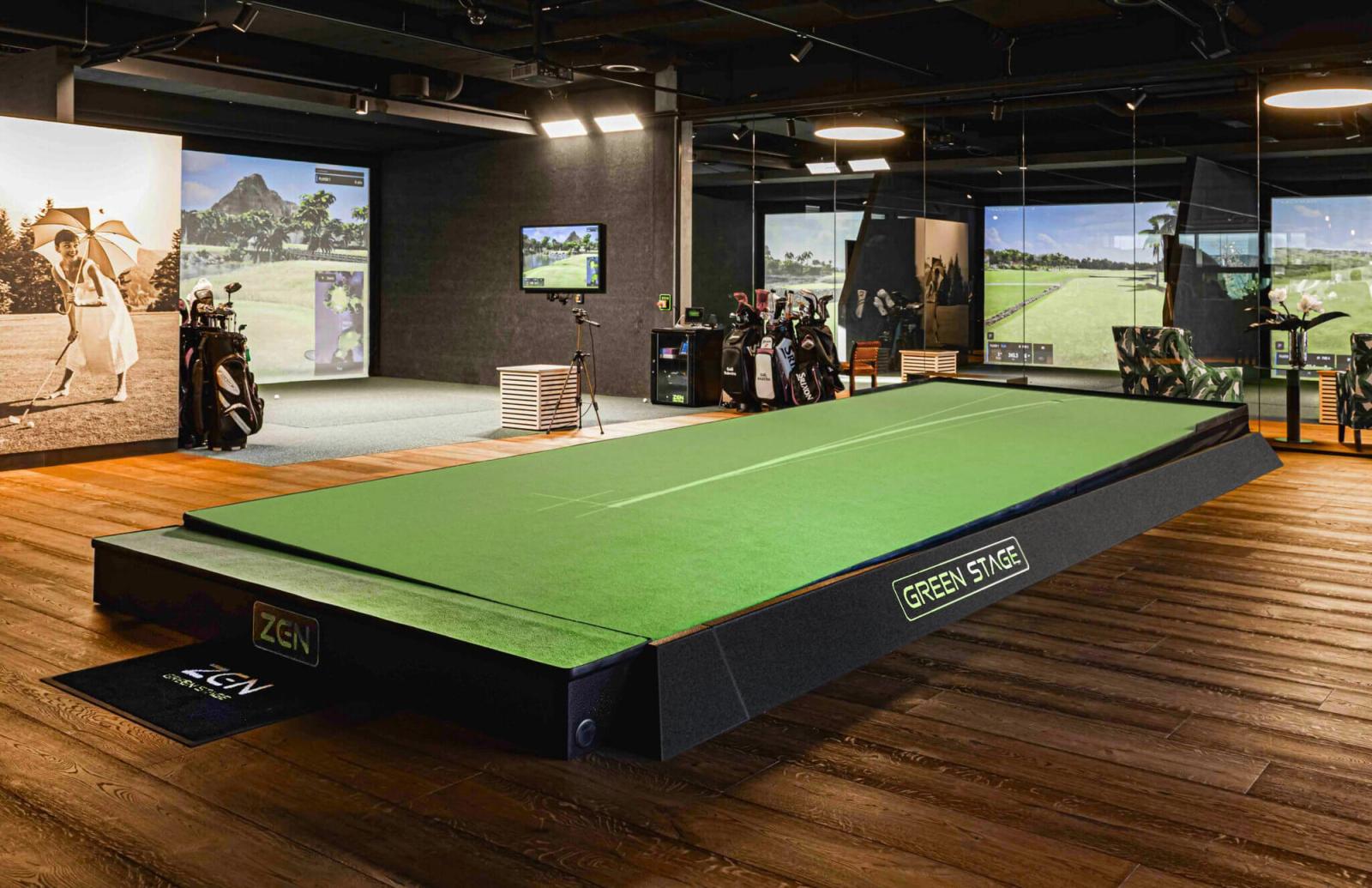 A modern indoor golf simulation room in London featuring a raised green putting stage, golf bags, and large screens displaying golf courses.