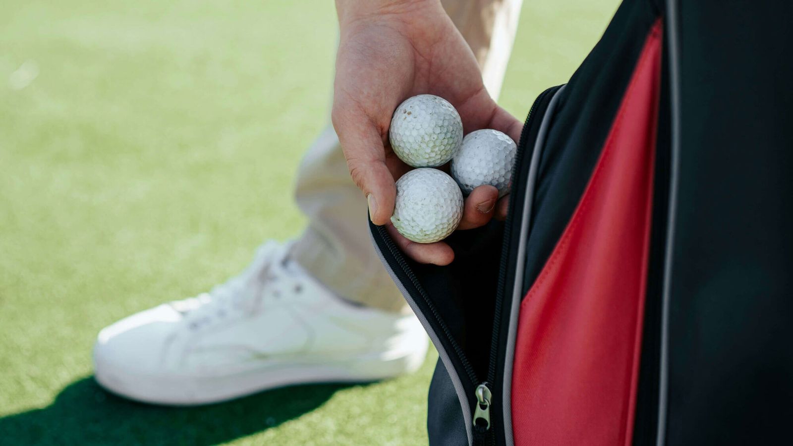 A person holds four golf balls in one hand while taking them out of a red and black bag, with part of a white sneaker visible on the green—capturing the essence of golf on the course.