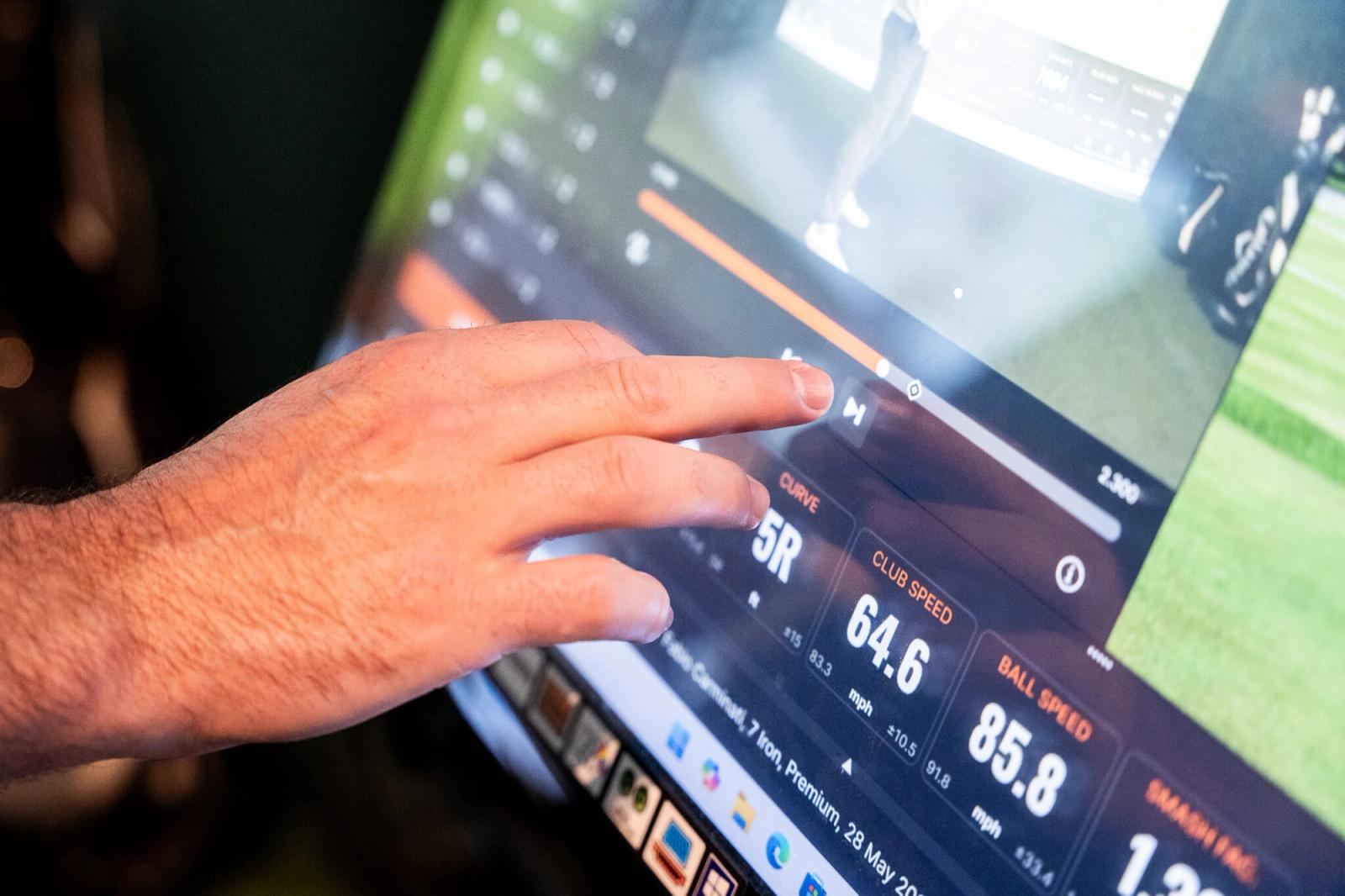 A person’s hand touches a digital screen displaying golf swing data, including club speed, curve, and ball speed—ideal for improving your game with personalized golf lessons or exploring exclusive golf memberships.
