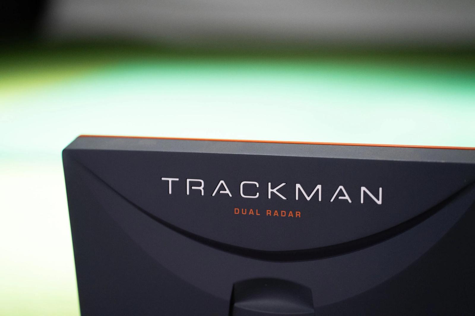 Close-up of a black device with the words TRACKMAN and DUAL RADAR in white and orange letters, set against a blurred green and yellow background—perfect technology for golf lessons or enhancing your golf memberships.