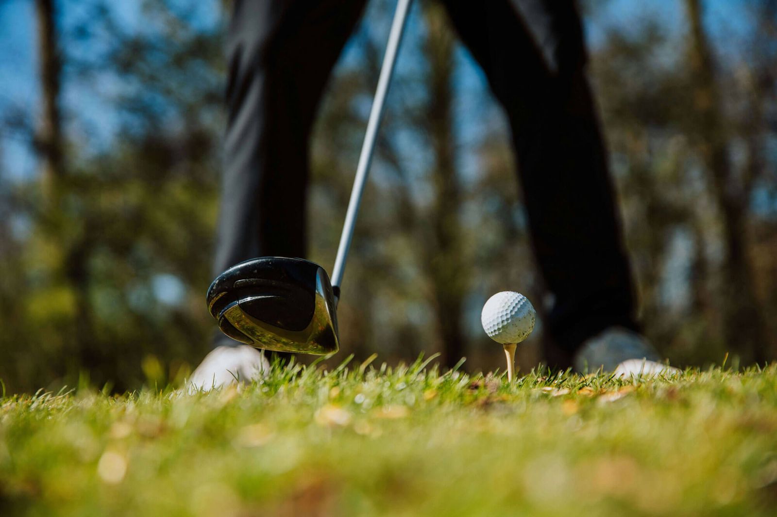 Close-up of a golfer preparing to hit a golf ball on a tee with a driver club, shown from a low angle on a grassy course with blurred trees in the background—perfect for those seeking premium golf memberships.