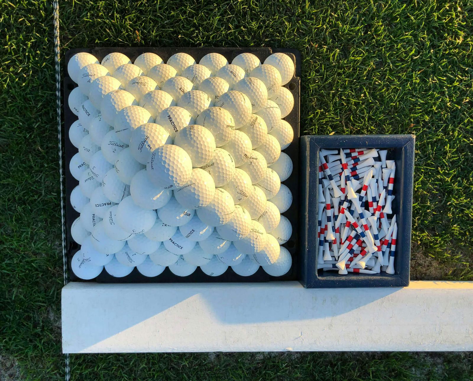A pyramid of stacked white golf balls sits on a black tray next to a box of red-striped tees, all placed on green grass—perfect for those seeking golf lessons or exploring golf memberships in London.