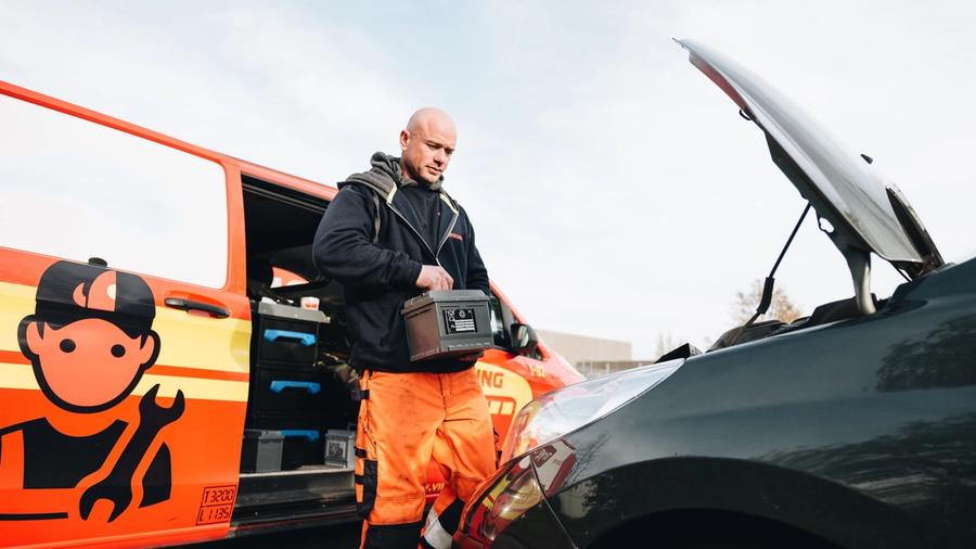 Mies oransseissa työhousuissa ja mustassa takissa seisoo avoimen konepellin vieressä pitäen kädessään auton akkua. Lähistölle on pysäköity mekaanikkologolla varustettu huoltoauto.