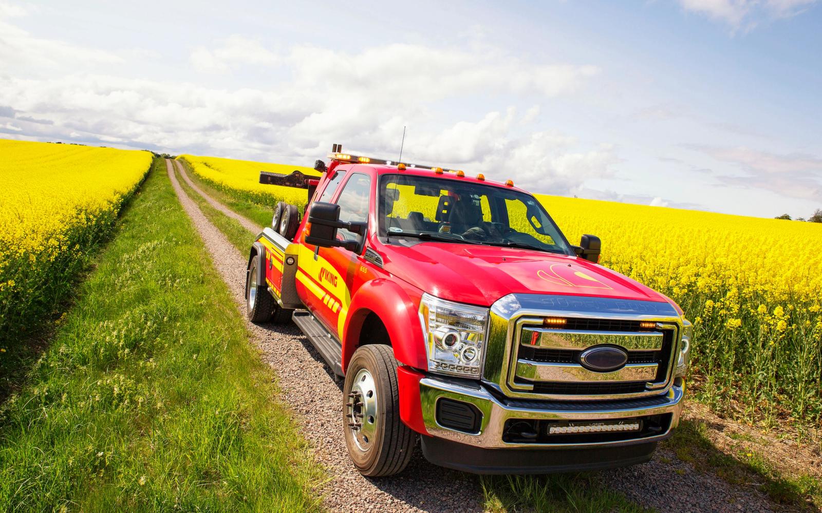 A red Viking tow truck is parked on a gravel road between fields of yellow flowering plants under a partly cloudy sky.
