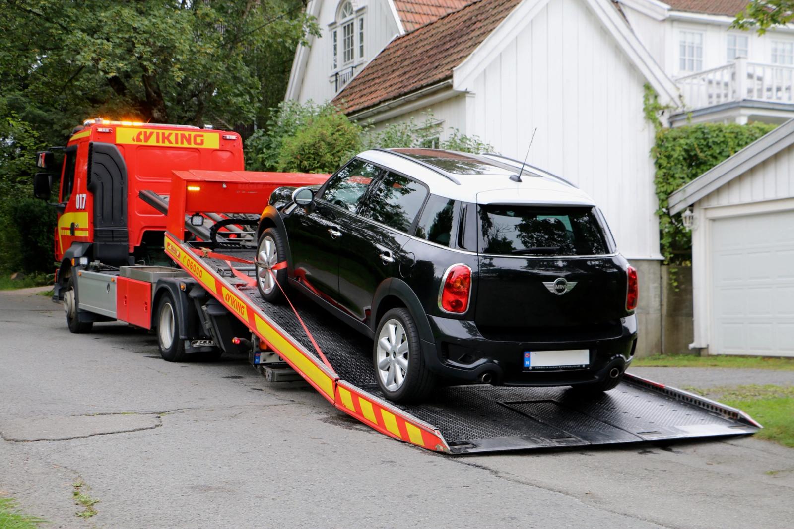 Mustavalkoinen auto lastataan hinausautoon asuinkadulla, jonka taustalla on valkoinen talo taustalla.
