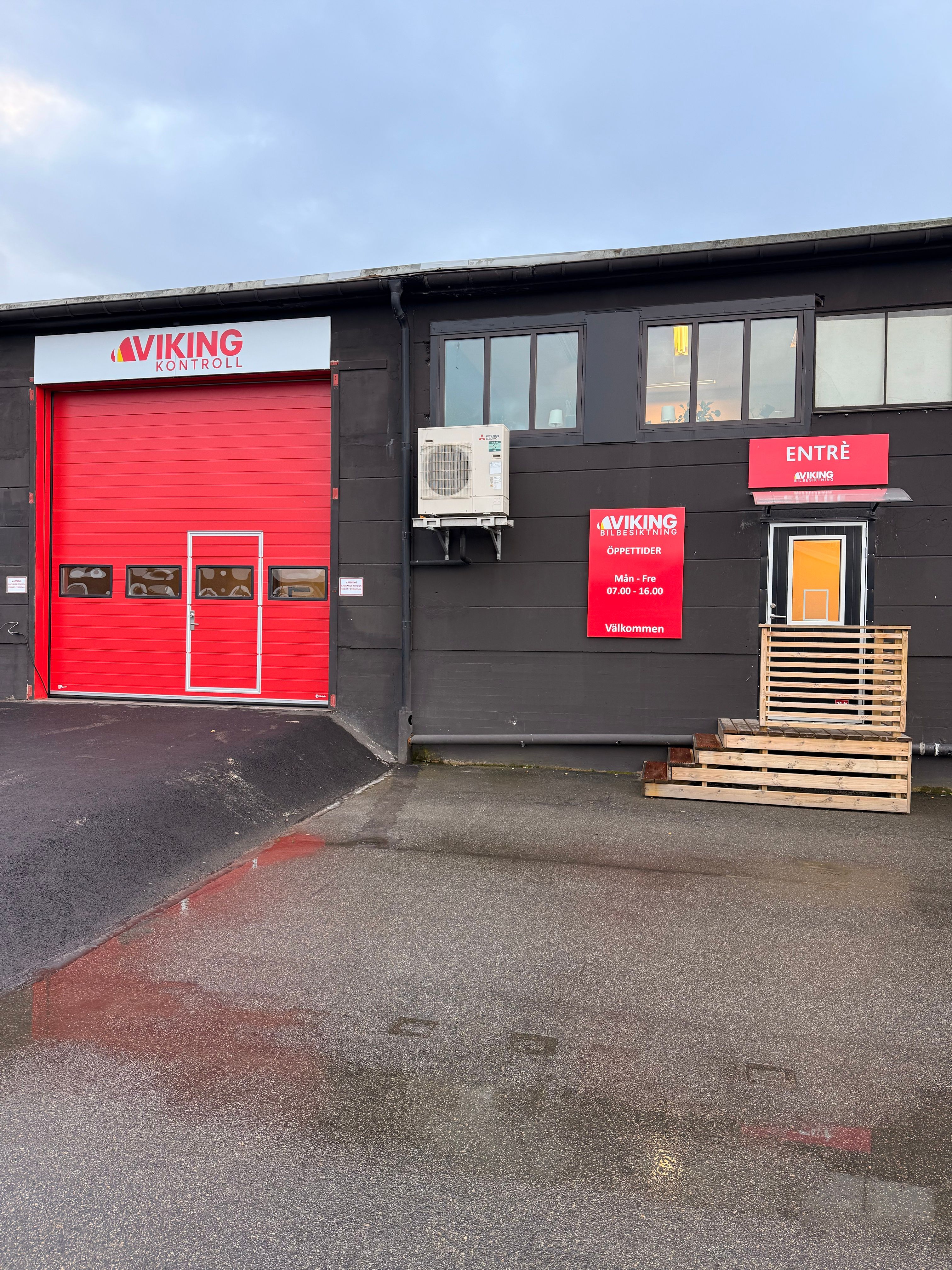 Utsidan av en fordonsbesiktningsstation från Viking Bilbesiktning med två röda garageportar, en ingång och en företagsbil parkerad till höger.