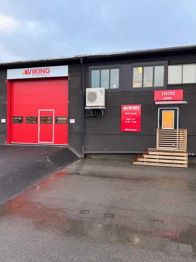 Utsidan av en fordonsbesiktningsstation från Viking Bilbesiktning med två röda garageportar, en ingång och en företagsbil parkerad till höger.