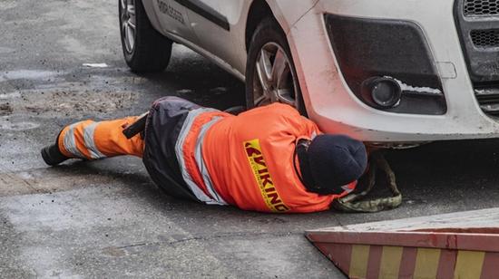 Oranssissa työasussa oleva henkilö makaa valkoisen ajoneuvon etuosan alla suorittamassa huoltoa tai tarkastusta.