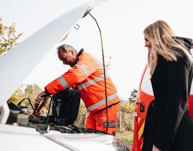 VIking työasuun pukeutunut hinausauton kuljettaja antaa apuvirtalaitteella käynnistysapua autolle naisen katsellessa.
